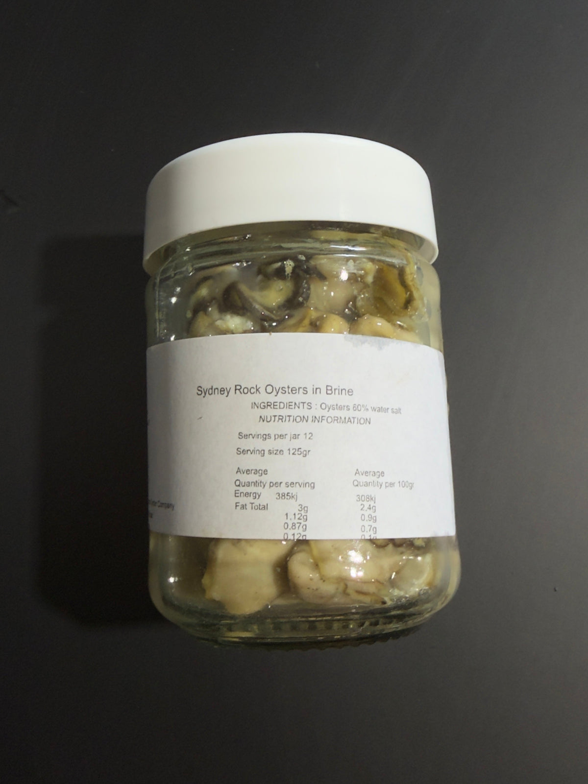 A clear glass jar of Sydney Rock Oysters in Brine with a white lid and white label, set against a dark gray background.