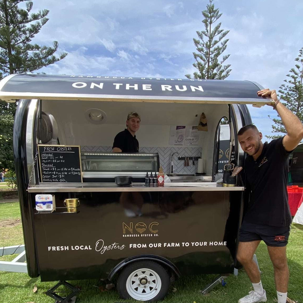 A black mobile food truck with a white canopy is parked on grass outdoors under a blue sky. A man in a black shirt and shorts stands beside the open serving window holding the canopy up, while another person is visible inside the truck.
