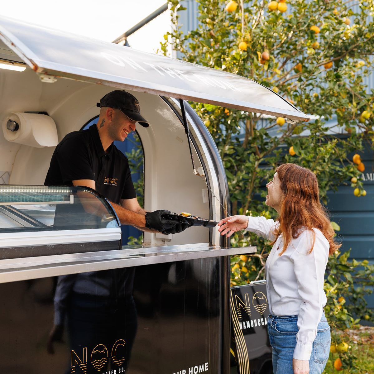 A mobile oyster food truck with an awning is parked outdoors on grass next to an orange tree. A man wearing a black polo shirt and cap serves food from the truck to a woman in a white long-sleeve shirt and blue jeans.