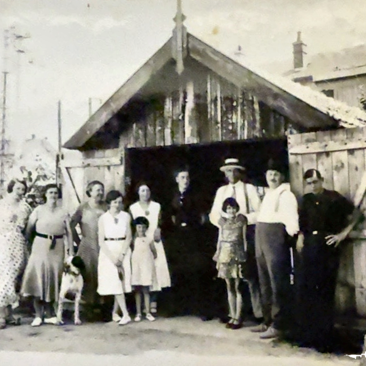 An aged black-and-white photograph showing a group of people standing in front of a wooden shed or barn with open doors. 