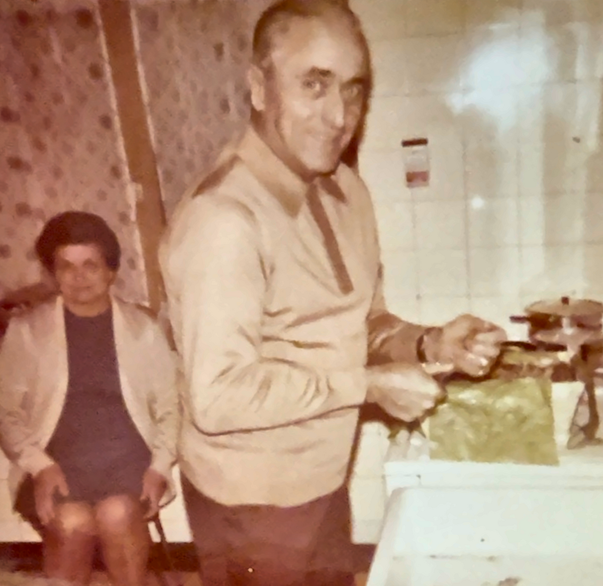 A vintage, warm-toned photograph showing a man in a beige sweater standing in a kitchen next to a sink, with a woman seated in the background. The photo has a white border and an aged, scanned-paper appearance.