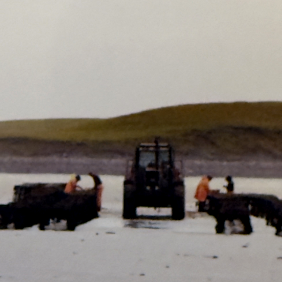 A wide outdoor scene showing a tractor and workers operating machinery on a flat, pale ground in front of a large grassy hill under an overcast sky.